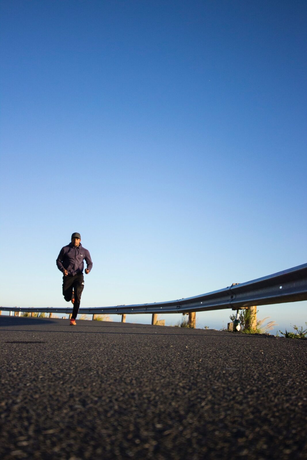 A person runs on an empty road near a metal crash barrier under a clear blue sky, wearing dark sports clothing and a hat—finding anger control through movement. The low-angle shot emphasises the vast expanse of sky overhead.