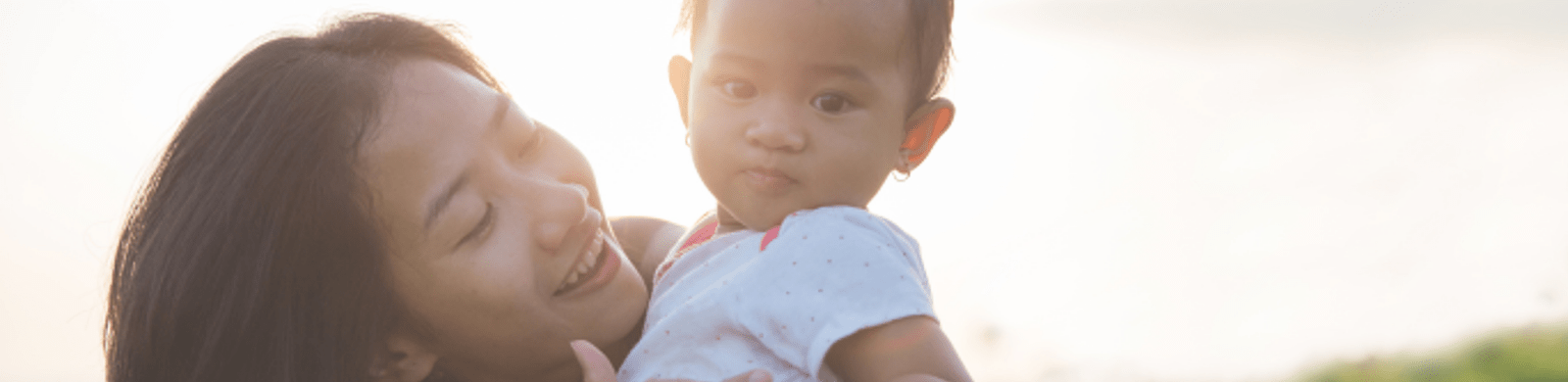 A smiling woman holds a baby outdoors, sunlight softly illuminating them from behind. The baby looks at the camera while the woman gazes at the baby affectionately—capturing a warm moment that parenting hacks can help create every day.