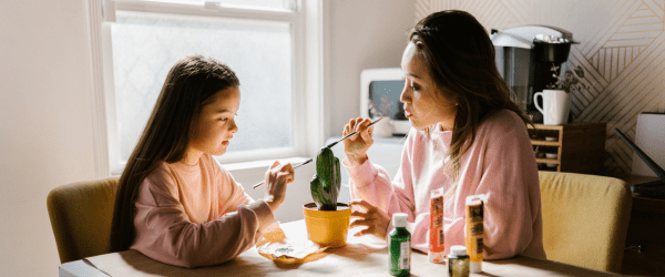 A woman and a young girl sit at a table painting a small flowerpot green together, connecting with your foster child through creativity, surrounded by paint bottles and sunlight streaming in from the window behind them.