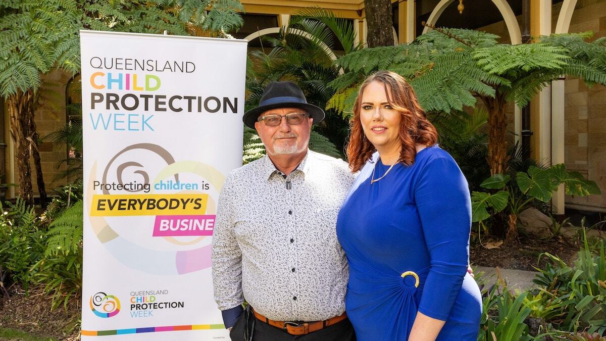 Deb and Steve stand outdoors beside a Queensland Child Protection Week banner. The man wears glasses, a hat, and a patterned shirt; the woman wears a blue dress. Green plants and an arched building are in the background.