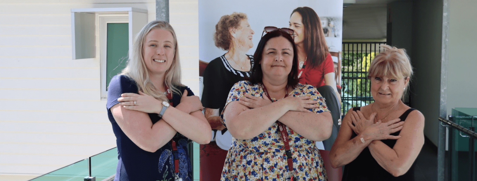 Three women standing side by side, smiling with arms crossed over their chests. They are indoors, with a light background and a poster behind them showing two women talking about women's homelessness.