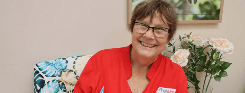 A smiling woman wearing glasses and a red shirt sits in an office chair with a floral pattern, embodying her nursing dream whilst on dialysis. Light pink roses and green leaves in a vase add charm beside her.