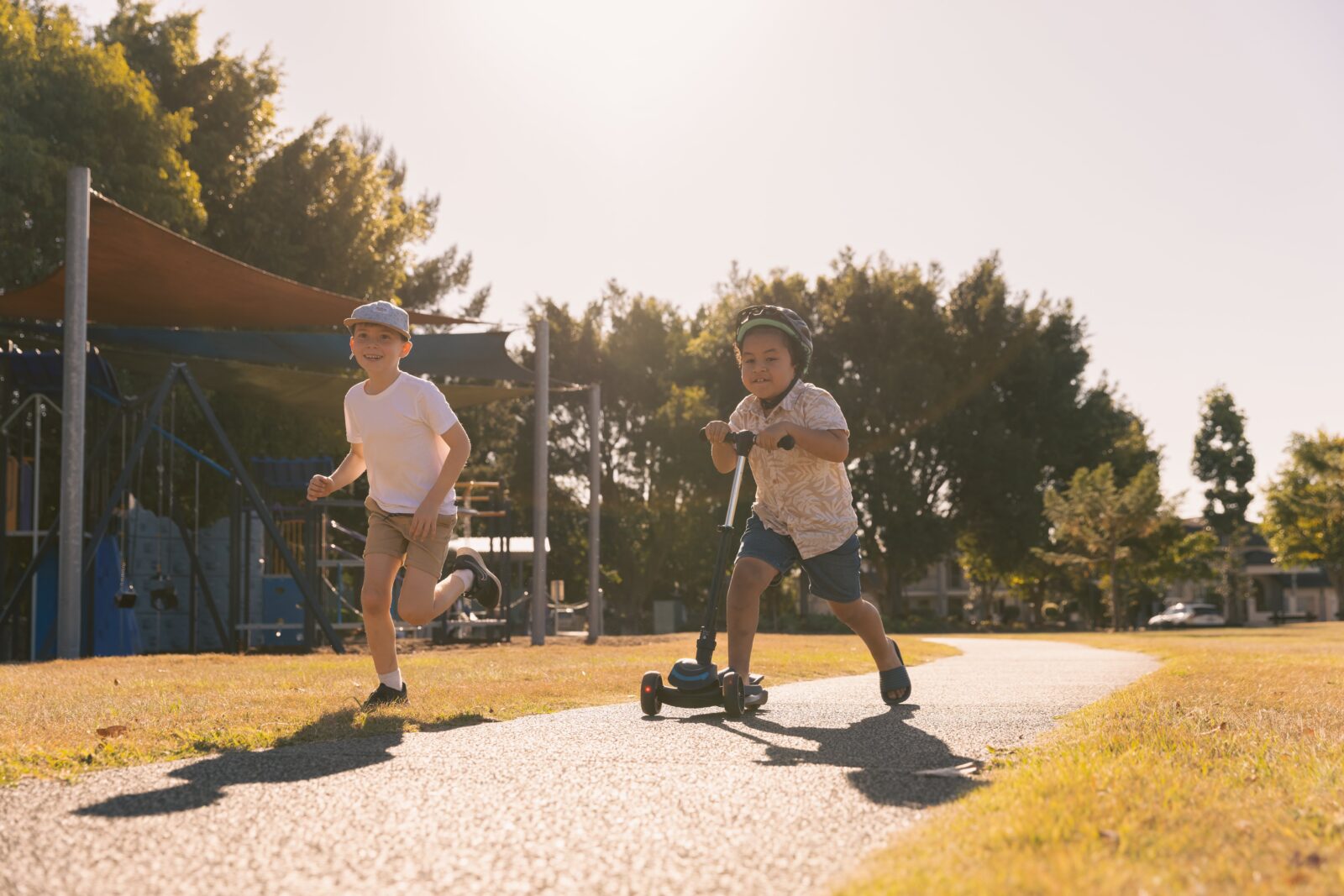 Two young children play outside on a sunny day; one rides a scooter while the other runs beside him near a playground—moments like these may answer the question, what is happiness? Both look joyful and full of energy as they enjoy their carefree afternoon.