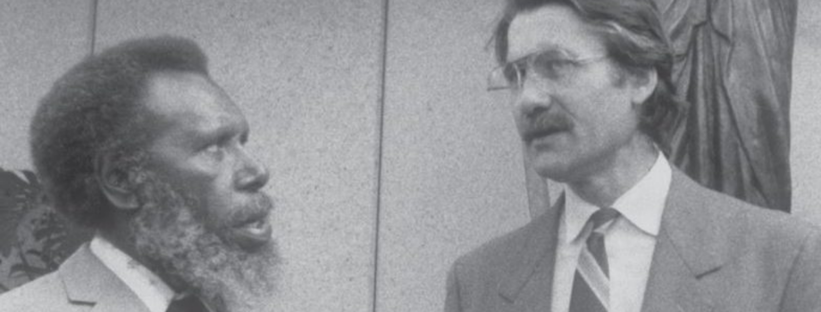 Two men in suits are engaged in conversation about the Mabo case. One has a beard and afro hairstyle, whilst the other has glasses and a moustache. They are standing in front of a plain wall.