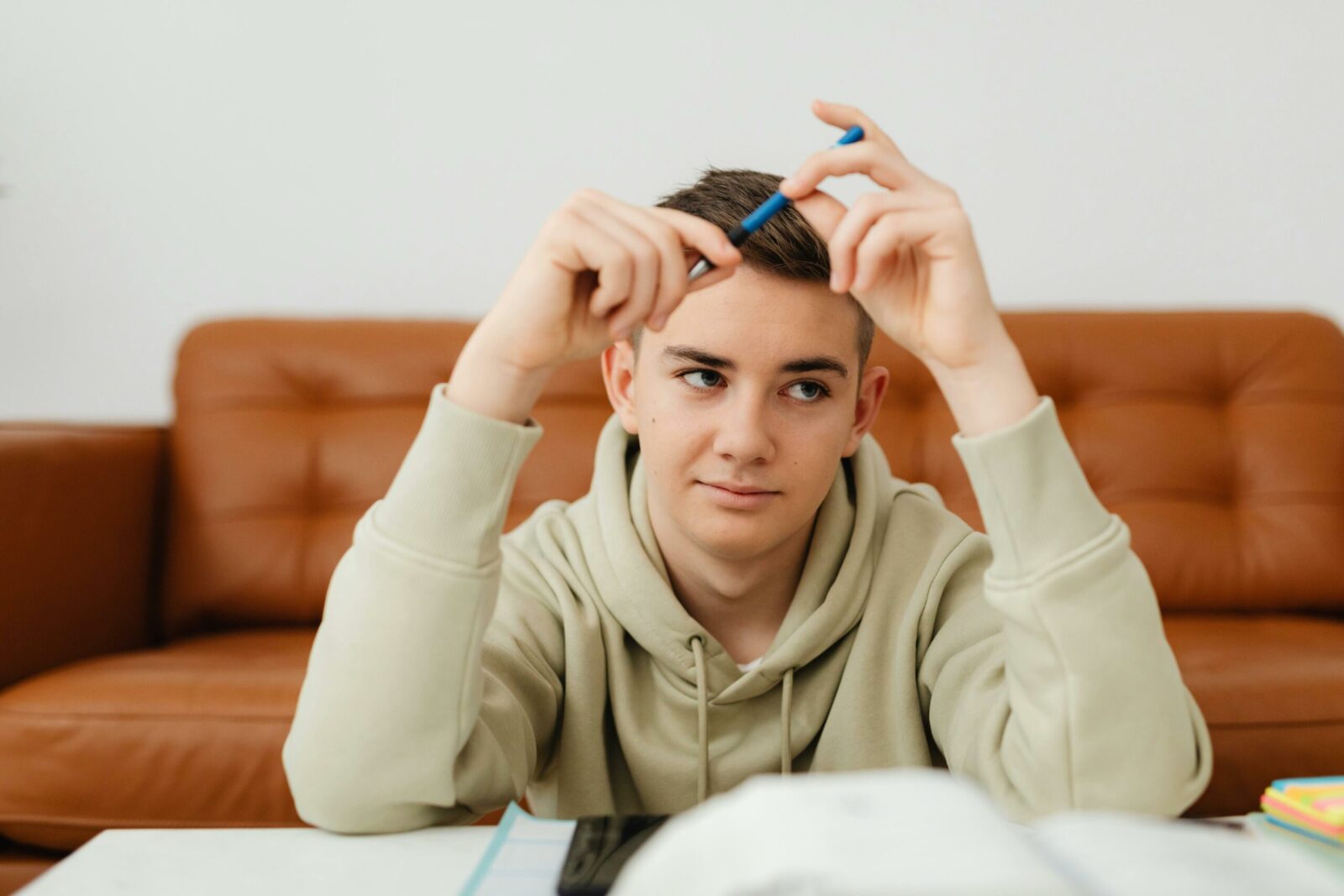 A teenage boy in a light hoodie sits at a table with books and pens, looking thoughtful while holding a pen near his face, deep in personal problem solving. A brown sofa and white wall are in the background.