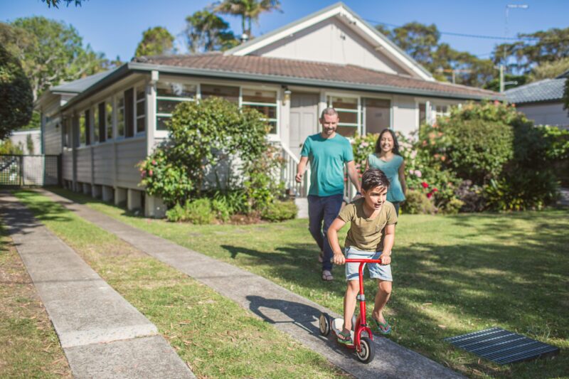 Family standing outside of their Queensland home with kid on scooter out the front on the driveway.