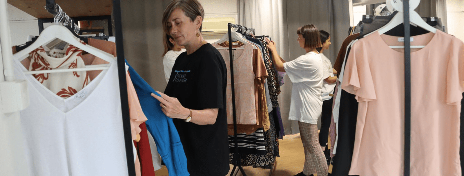 Two women browse through rails of colourful clothing in a well-lit Thread Together clothing hub, with various tops and dresses hanging on display.