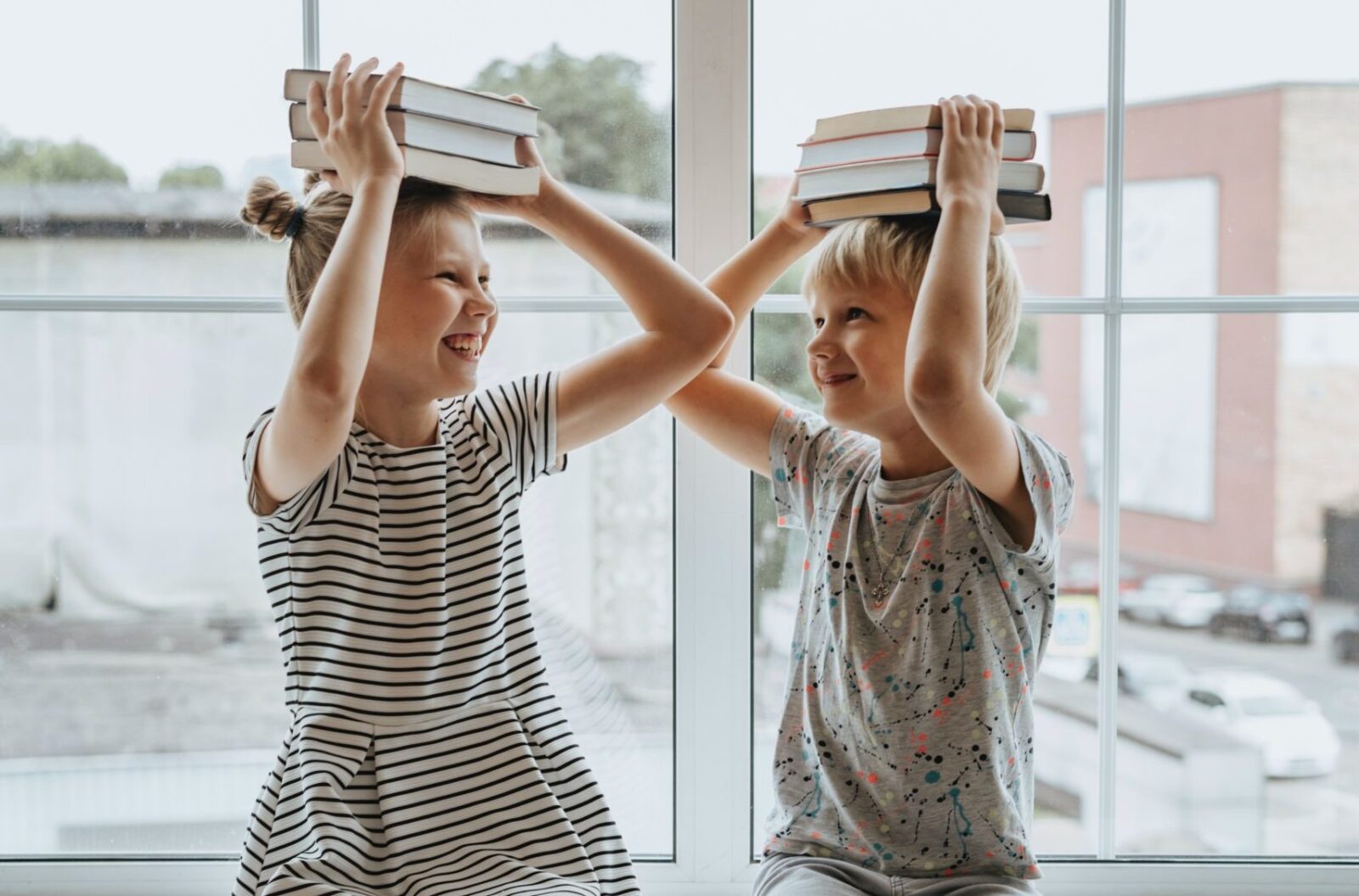 Two smiling children sit by a window, balancing stacks of books on their heads. As one child starts prep, they face each other and laugh, with bright daylight streaming in behind them.