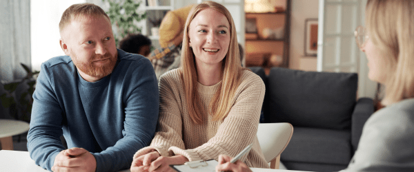 A smiling couple sit at a table facing a person with a clipboard, engaged in conversation about questions like, “Can I choose my foster child?” in a cosy, well-lit living room.