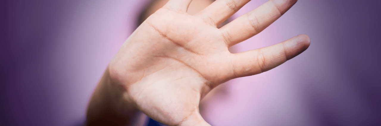 A close-up of a hand held up in front of the camera, palm facing outward in a gesture symbolising resistance to domestic and family violence, with a blurred background in shades of purple and pink.