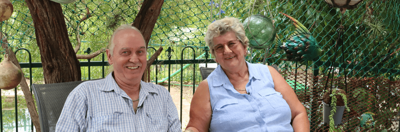 Foster carers Mark and Jenny sit smiling outdoors on a patio, surrounded by plants, green netting, and hanging ornaments, with trees visible in the background.