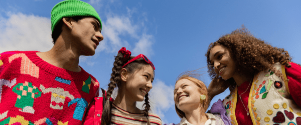 Four young people stand close together outdoors, smiling and talking under a blue sky. Wearing colourful, vibrant clothing and embracing sustainable choices, they appear happy, enjoying each other's company.