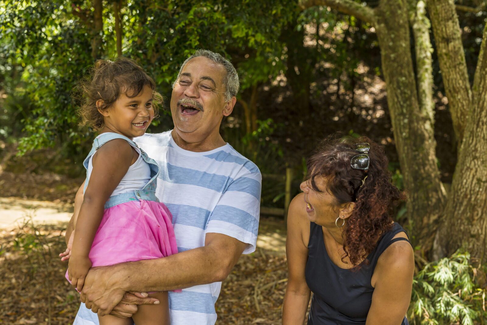 An older man holding a young girl in his arms, both smiling and laughing, while a woman beside them laughs as well. Surrounded by trees and greenery, they share a joyful moment outdoors, strengthened by cultural support.
