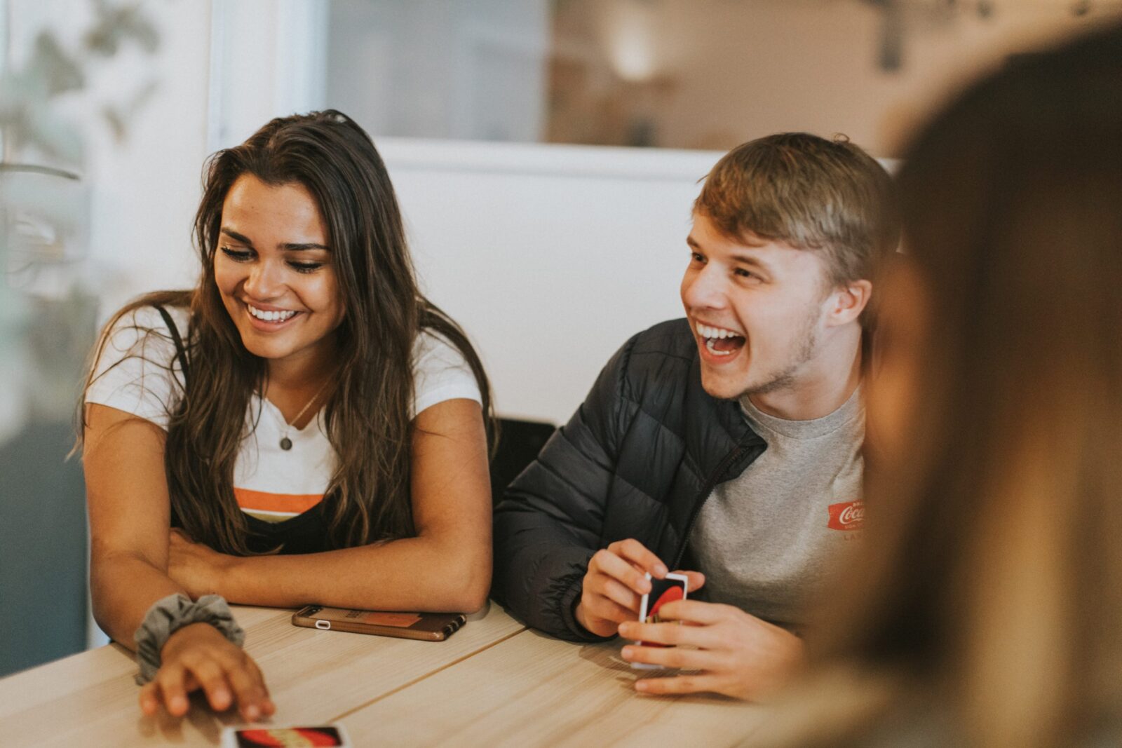 Two young adults sit at a table, smiling and laughing as they play a card game—capturing the joyful moment as a foster child turns 18. One reaches for a card whilst both enjoy the fun, with a blurred figure in the foreground.