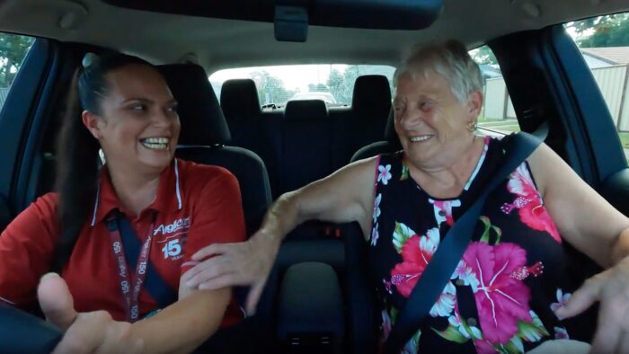 A woman in a red shirt and a name badge is driving a car, smiling and laughing with an elderly woman in a floral dress sitting in the passenger seat. Both are wearing seat belts and appear happy.