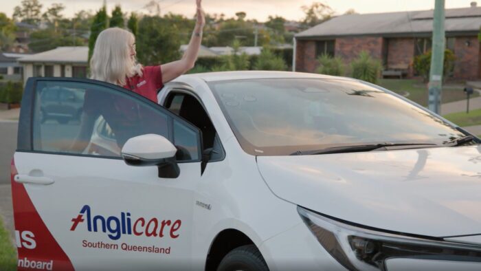A person with fair hair stands beside an Anglicare Southern Queensland car, waving whilst holding the driver’s door open on a suburban street.