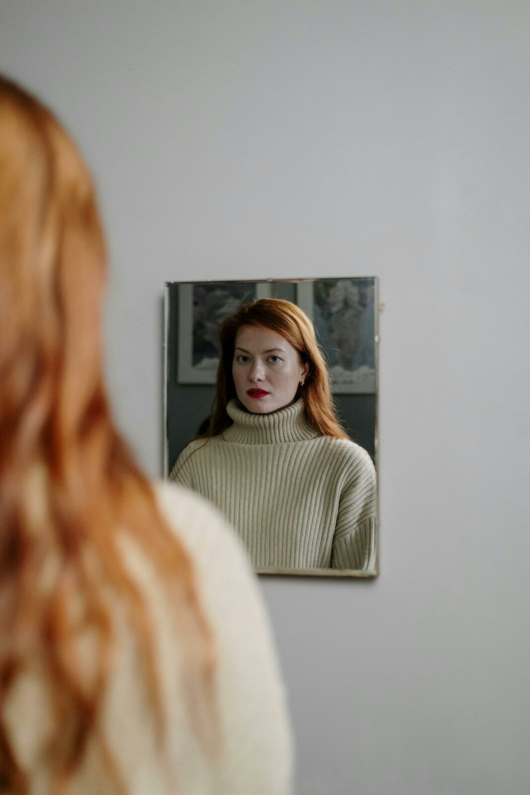 A woman with long red hair wearing a cream polo-neck jumper looks at her reflection in a wall-mounted mirror, contemplating her core beliefs, with her back slightly visible in the foreground.