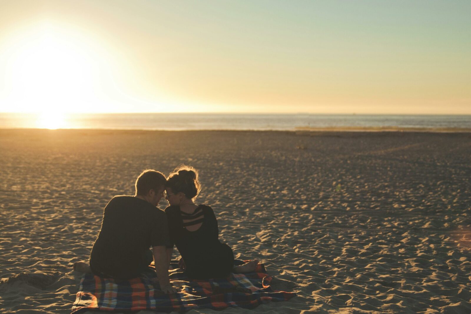 A couple sits closely together on a blanket at the beach, facing the sea as the sun sets, casting a warm golden glow over the sand—capturing one of the beautiful stages of a relationship.