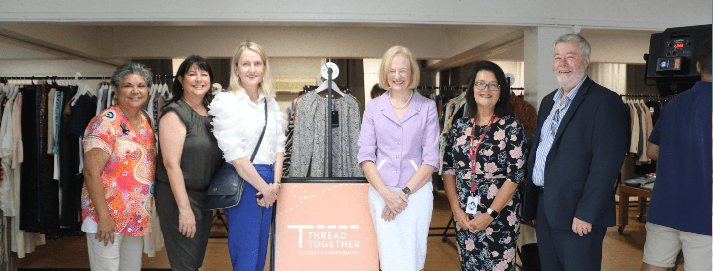 Seven people stand smiling indoors behind a rail of clothes and a sign that reads Thread Together Clothing Communities, highlighting their vibrant clothing hub. Clothes rails are visible in the background.
