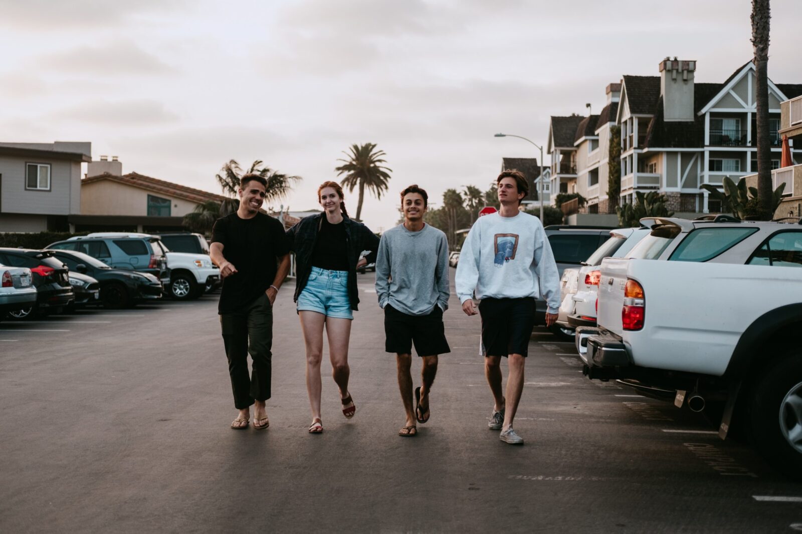 Four young adults walk barefoot down a street lined with parked cars and palm trees, smiling and talking together as they enjoy The Home Stretch. Houses and a cloudy sky are visible in the background.