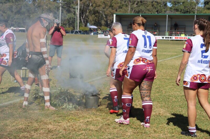 First Nations rugby players standing around on a football field during a smoking ceremony at the Frank Fisher Cup