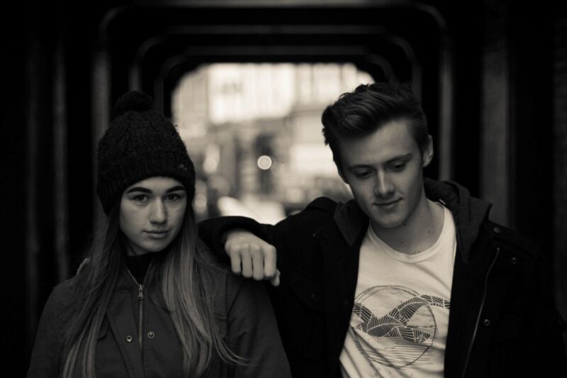 portrait of young man and women in city at night. She is wearing a beanie and he has a leather jacket on.