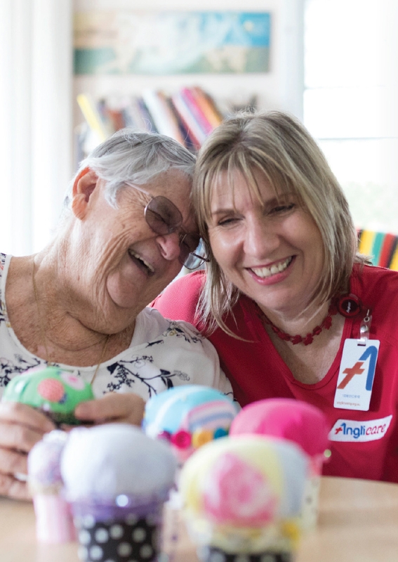 Two women, one elderly and one middle-aged, smile and lean close together at a table of colourful craft items. The younger woman, wearing a name badge and red shirt, helps assemble annual reports amid books and soft light in the background.