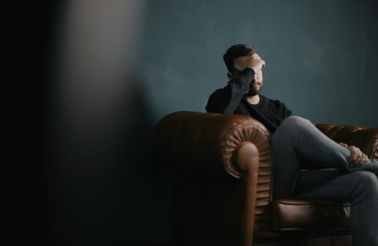A person sits on a brown leather sofa with legs crossed, resting their head in one hand, appearing stressed or deep in thought, anxiety evident against a dark, plain background.