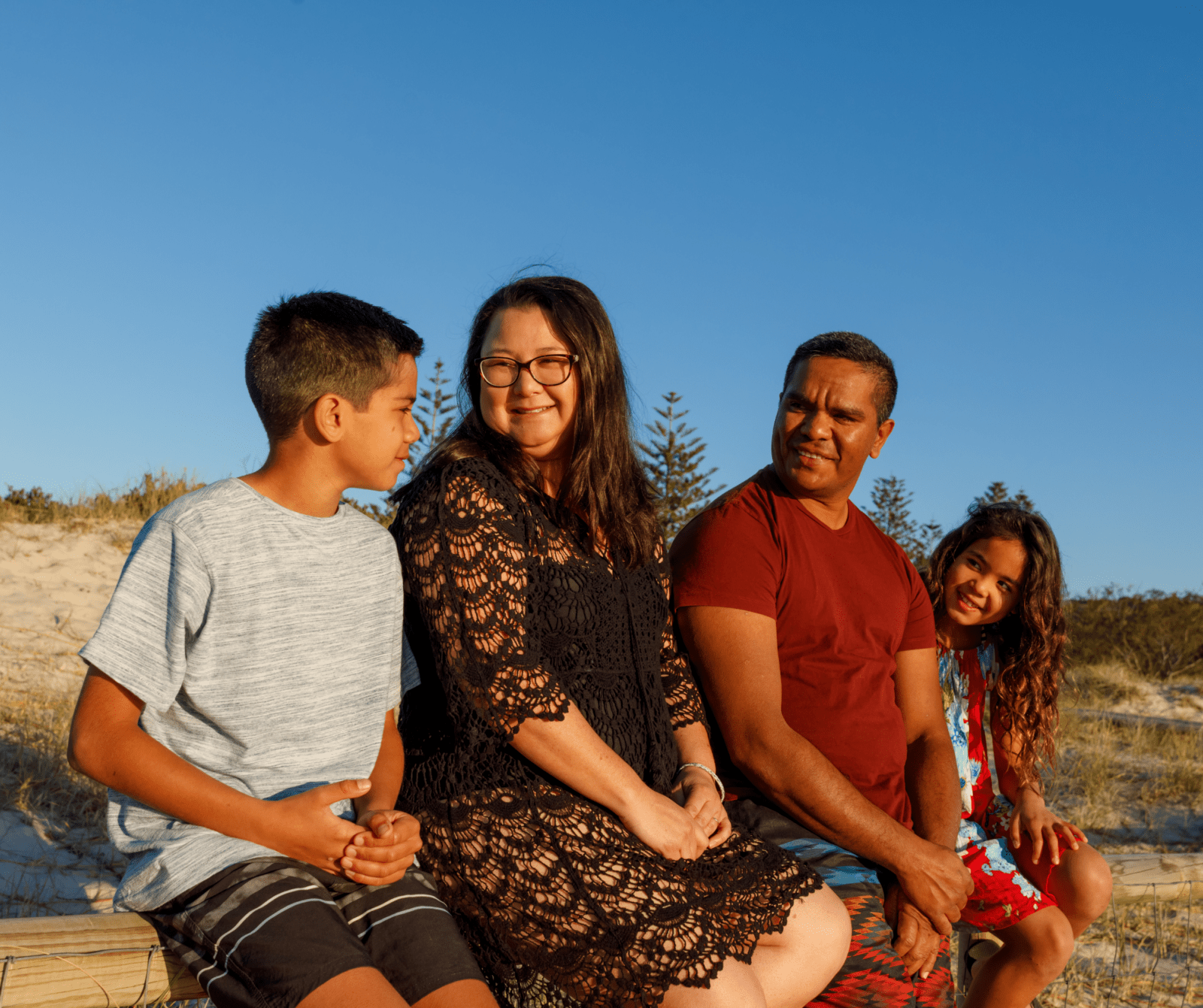 A blended family of four sits on a wooden bench at the beach, smiling and enjoying a sunny day. Two adults are in the middle with two children on either side, framed by sand dunes and a bright blue sky.