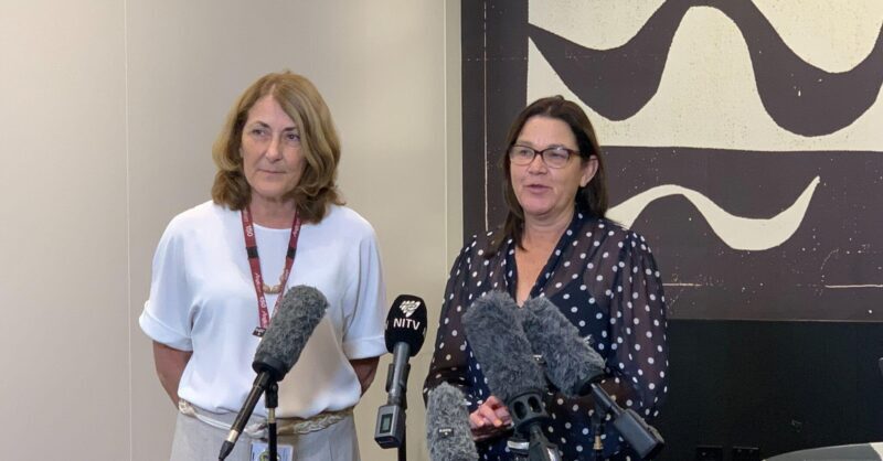 Two women stand in front of microphones at a press event focused on social justice. One wears a white blouse and lanyard, the other wears glasses and a dark polka dot blouse. Abstract art hangs on the wall behind them.