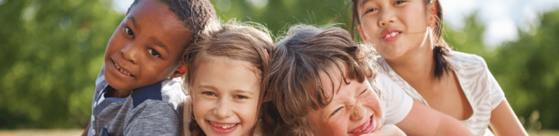 Four children smiling and hugging each other whilst playing outdoors on a sunny day, with green trees in the background, celebrating the spirit of Human Rights Day through friendship and unity.