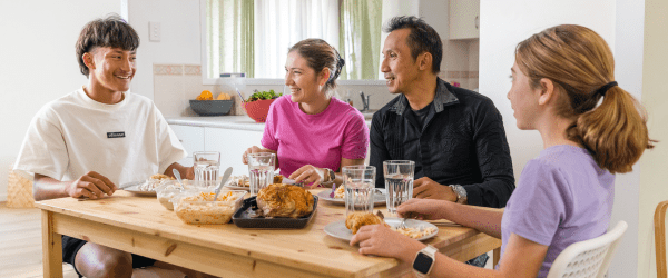 A family of four sits around a wooden table, smiling and enjoying a meal together with roast chicken, salad, and glasses of water in a bright, modern kitchen during National Families’ Week.