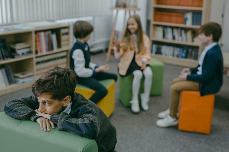 A boy sits alone on a green seat, looking sad, possibly experiencing bullying, whilst three other children interact together in the background of a library or classroom.